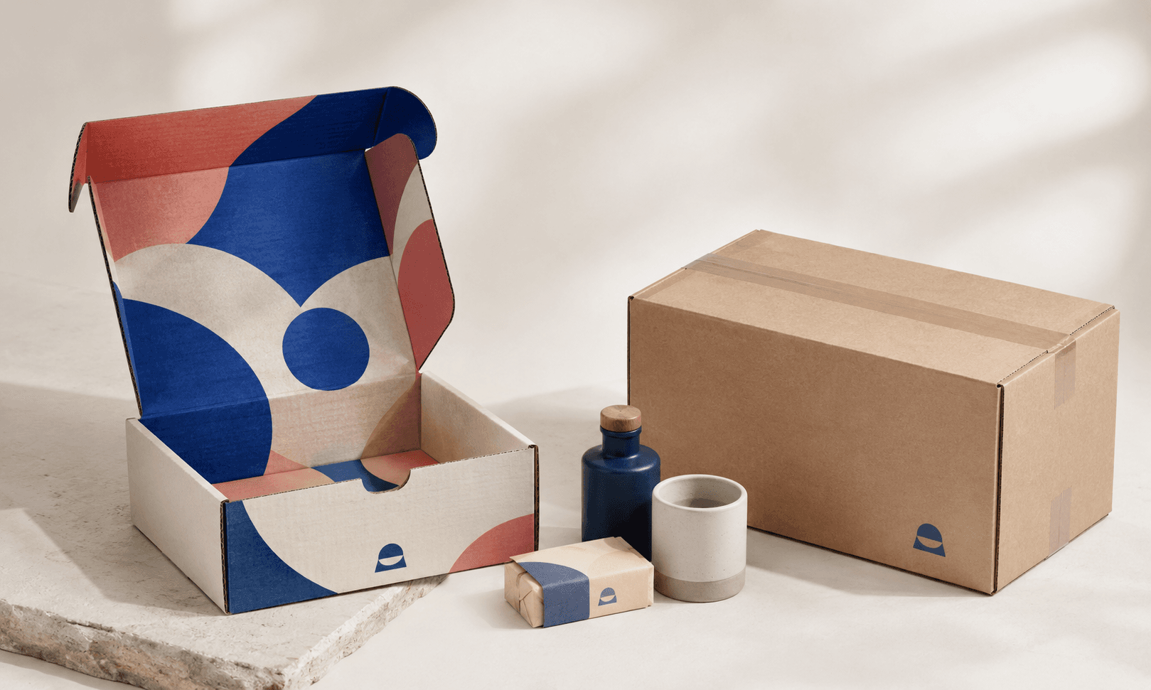 Branded corrugated mailer beside an H-taped shipping carton with compact ecommerce products arranged between them.